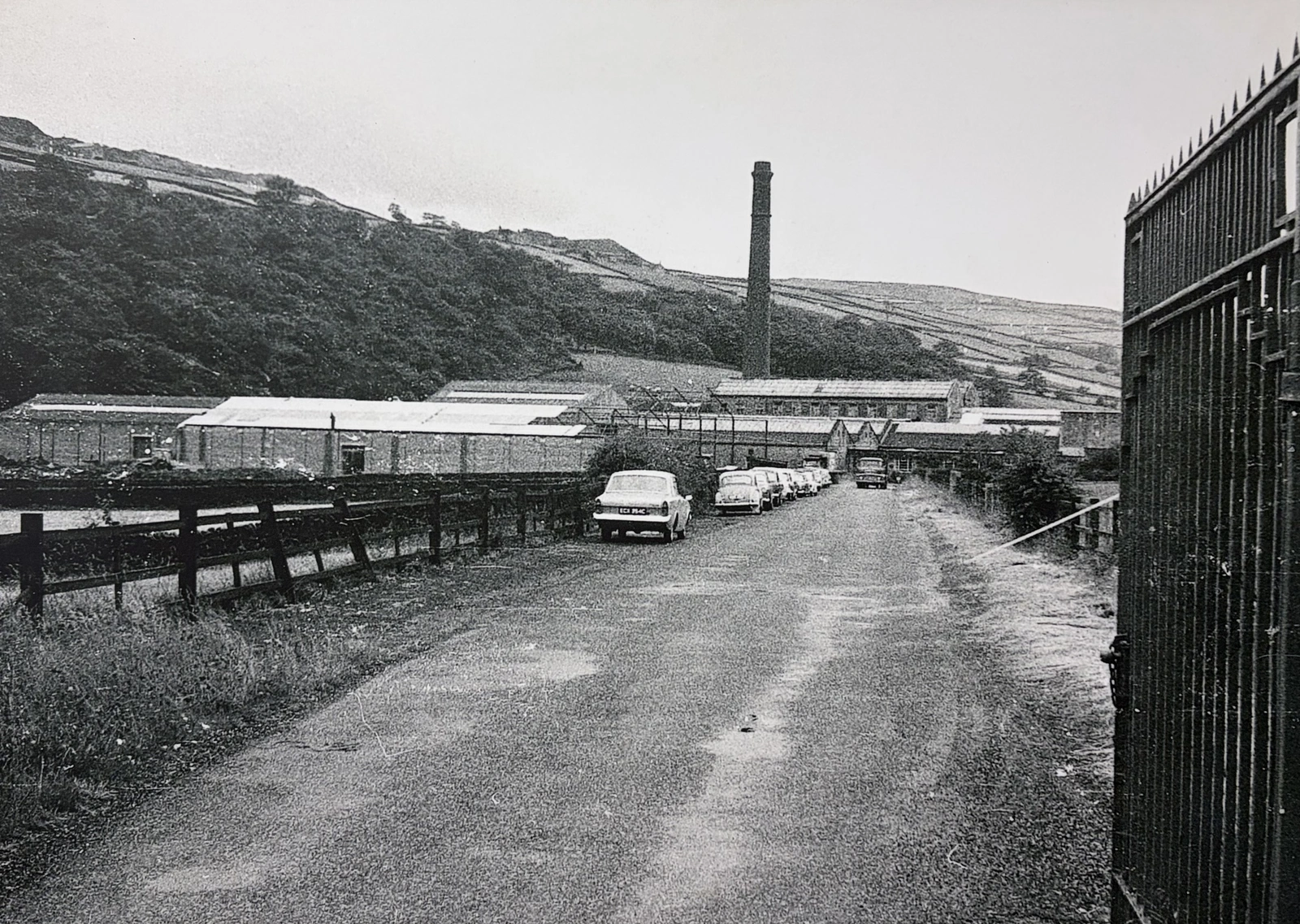Black-and-white archive photograph of mill buildings, a chimney and parked cars beneath the hillside